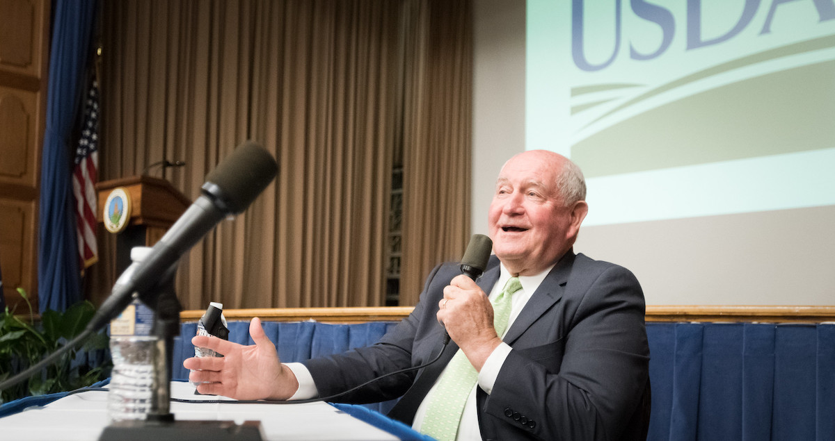 Sonny Perdue sits at a table and speaks into a microphone.