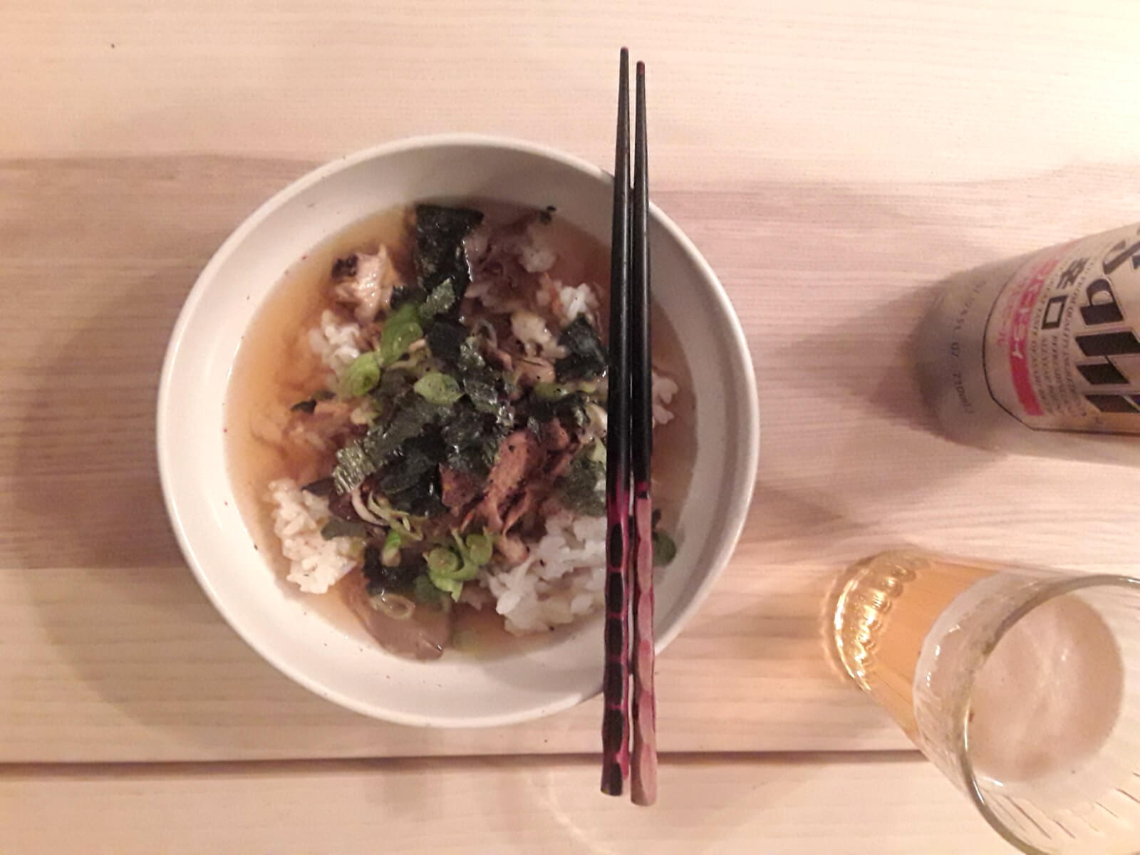 A bowl filled with dashi, rice, seaweed, and other garnishes sits on a table. A pair of chopsticks are on top of the bowl. Next to the bowl is a can of Asahi beer and a glass filled with the beer. (April 2020)