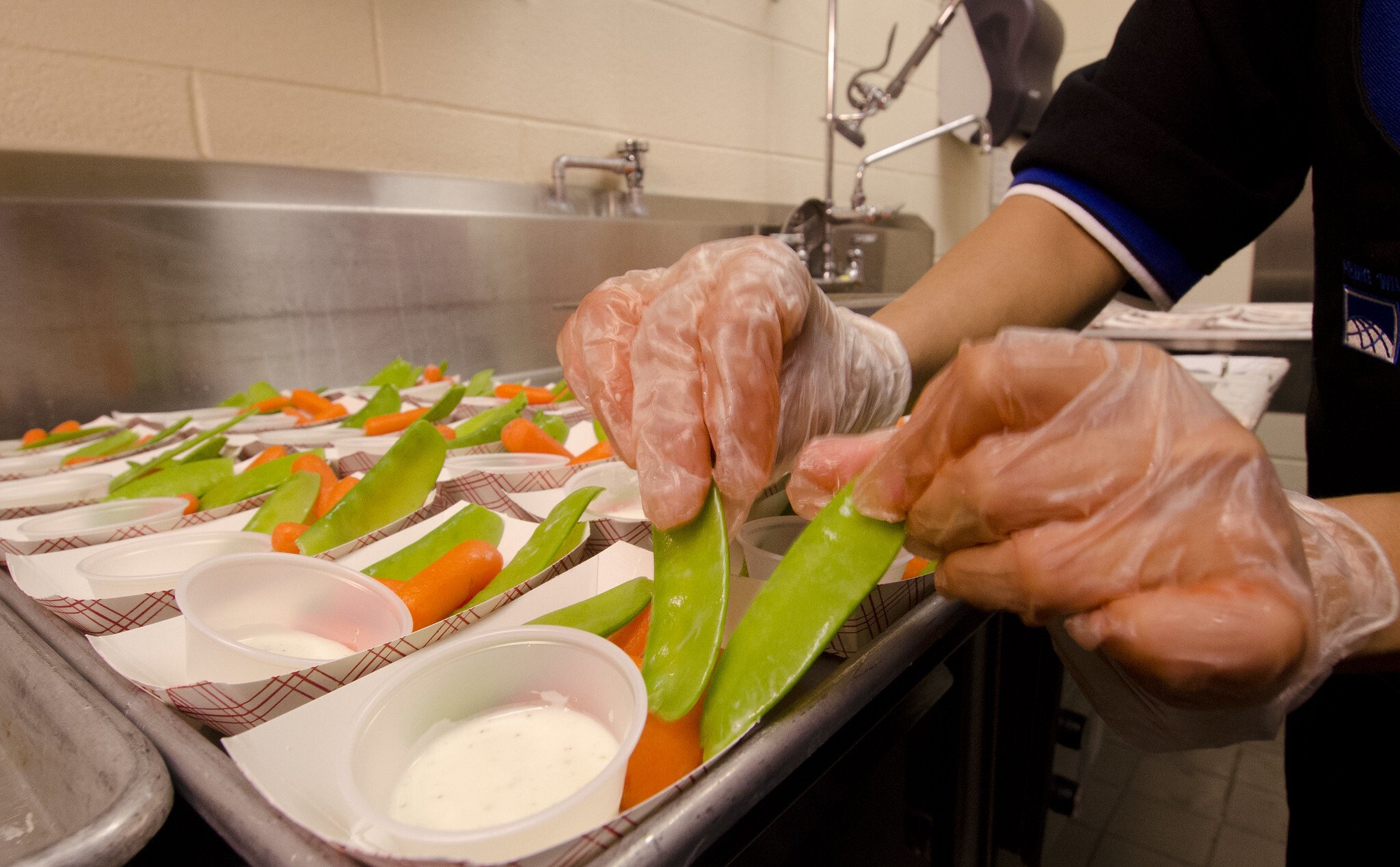 Two hands place snow peas on a school lunch tray with carrots and ranch dressing on the side. (April 2020)