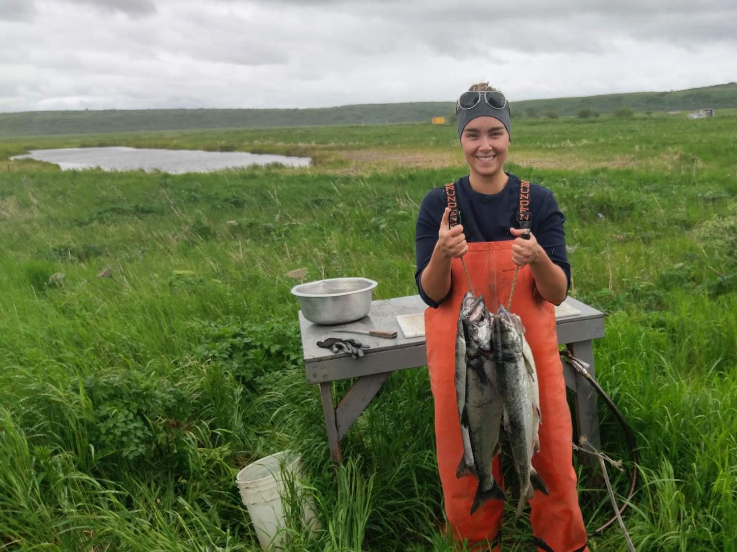Jamie O'Connor holds up fish in a grass field, Bristol Bay May 2020