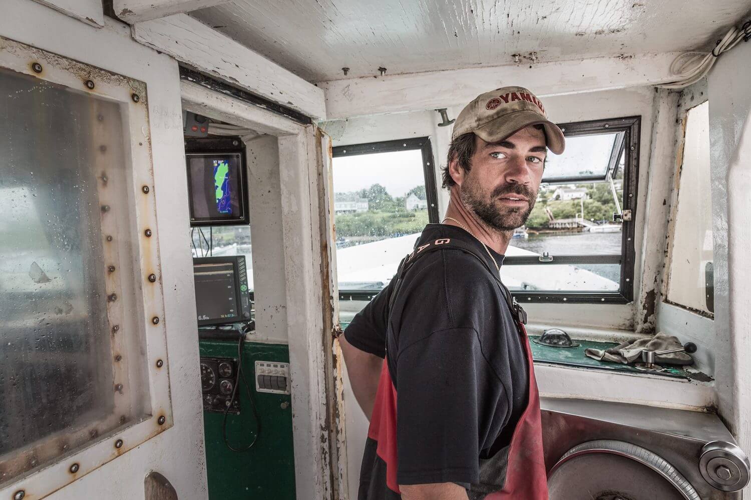 Herman Coombs, a lobster fisher in Orrs Island, Maine wears a a black t-shirt and looks back at the camera while on his fishing boat. (June 2020)