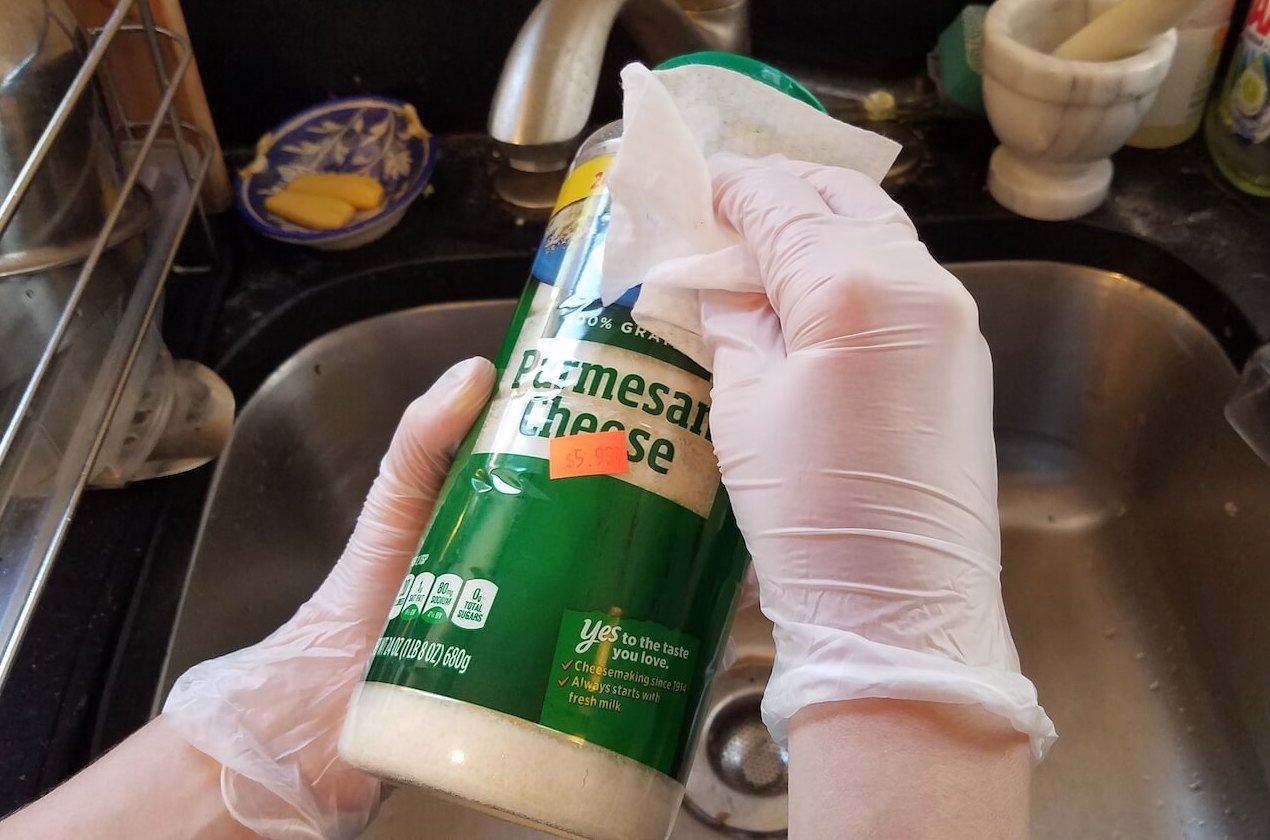 Two hands with gloves on wipes down a bottle of parmesan cheese as a safety precaution. (June 2020)