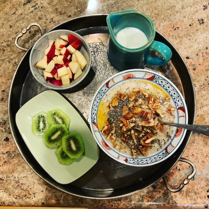 An ornate metal tray arranged with two bowls of fruit, a blue pitcher of warm milk, and a bowl of oatmeal topped with nuts, chia seeds, and honey. August 2020