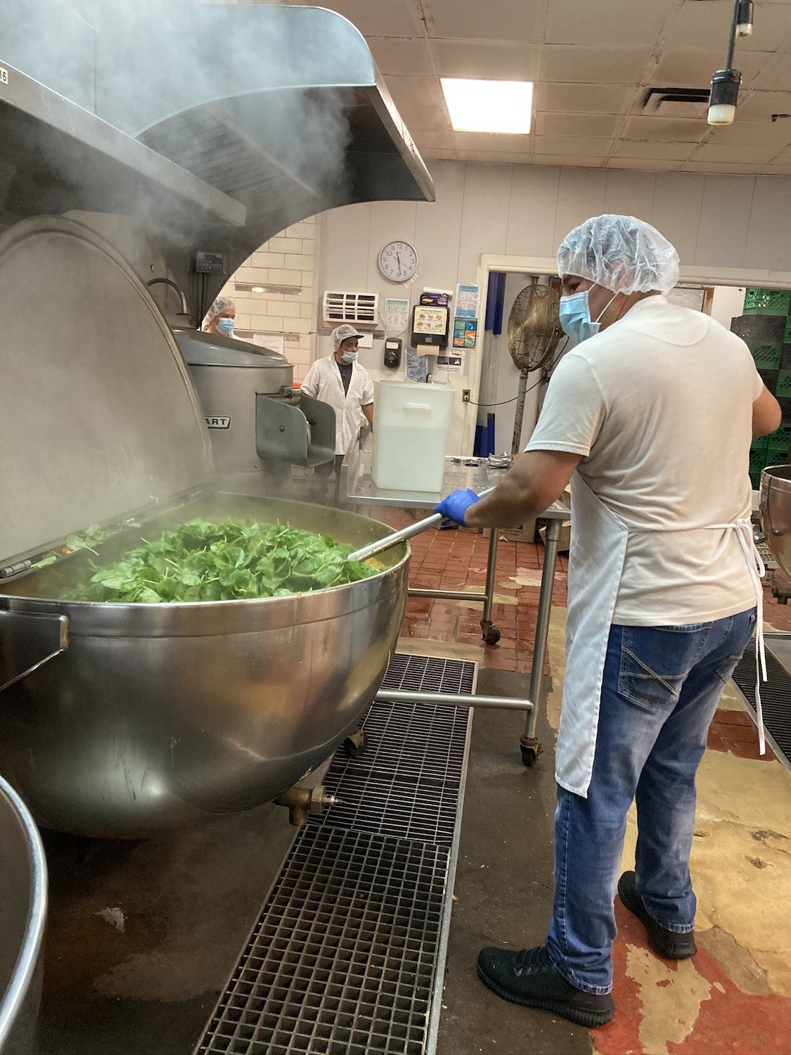 A worker cooks spinach in PPE in a large vat. October 2020