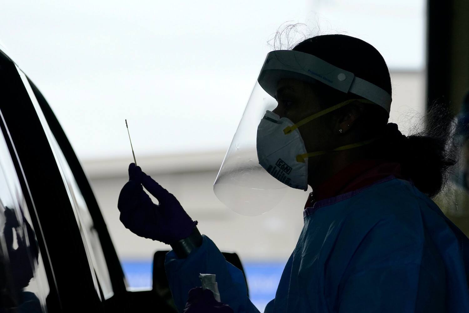 A woman wearing PPE holds a nasal swab up to a car for Covid-19 testing. October 2020