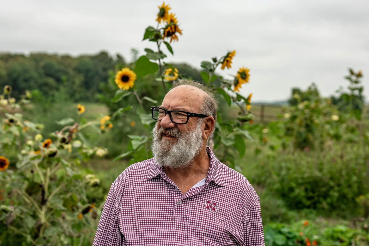 A man standing wearing glasses and a red plaid shirt in front of sunflowers. October 2020