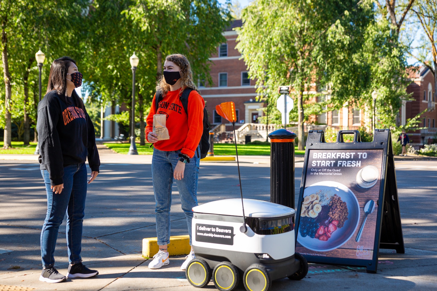 A delivery robot, students, and breakfast sign on a curb at Oregon State University. November 2020
