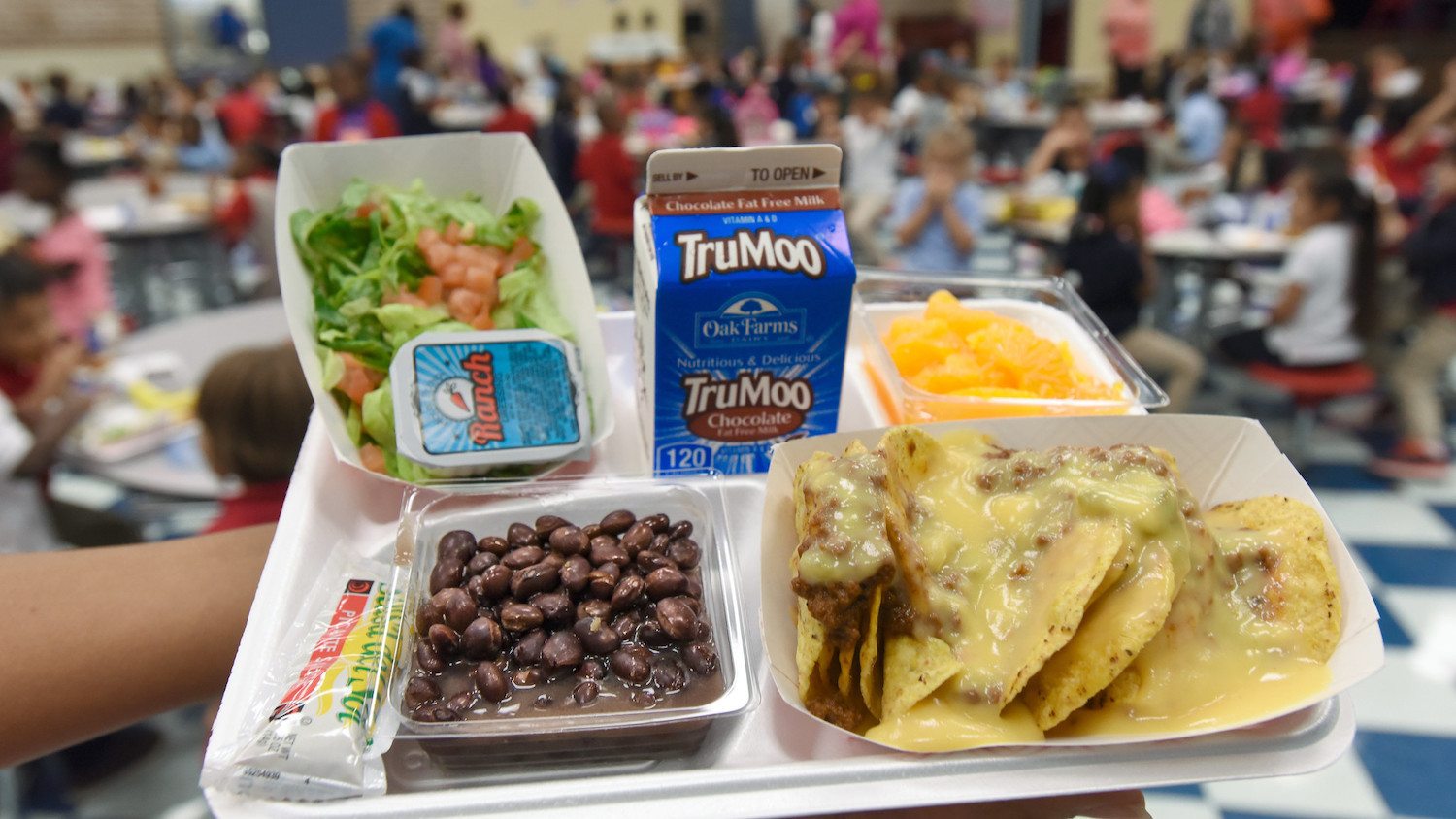A school lunch tray carried to a cafeteria table with nachos, beans, salad, fruit, and chocolate milk. November 2020
