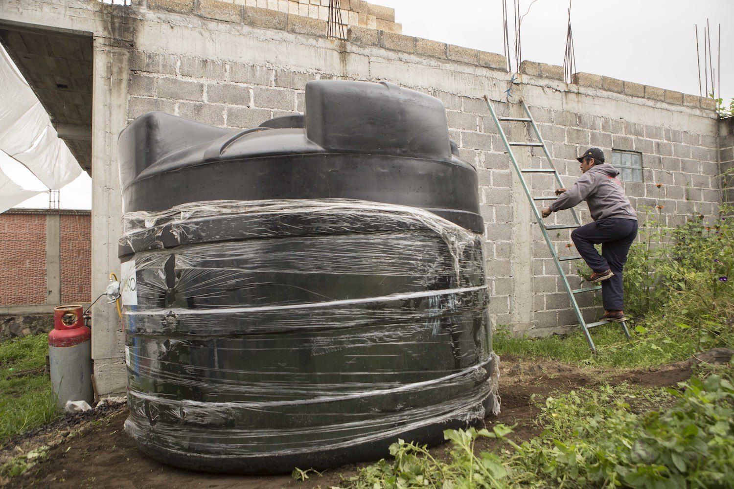 In this Sept. 3, 2016 photo, Edgar Serralde, a lettuce farmer in Mexico City’s borough Xochimilco, uses a ladder to climb on to his rooftop the day a large water cistern, left, was delivered to his home.