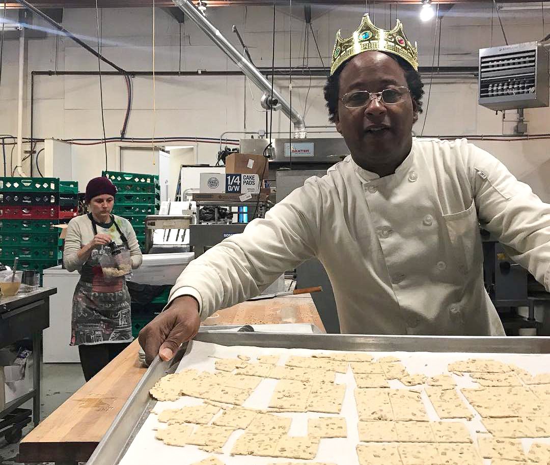 Jovani Prince wears a crown while holding a baking sheet in the kitchen of his gluten-free crackers towards the camera. A baker in the foreground is carrying supplies. January 2021