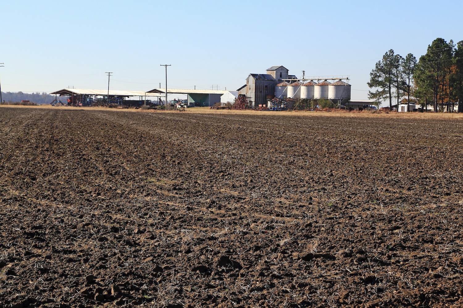 Large black land farm in Eastern Arkansas with plowed field laying fallow. Seed and grain silos in background with sheds and farm equipment.