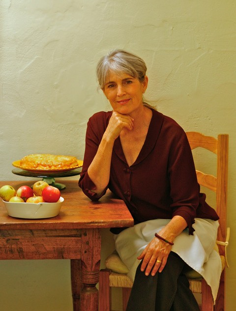 Vertical portrait of Deborah Madison sitting at a table with apples and a pie. June 2021