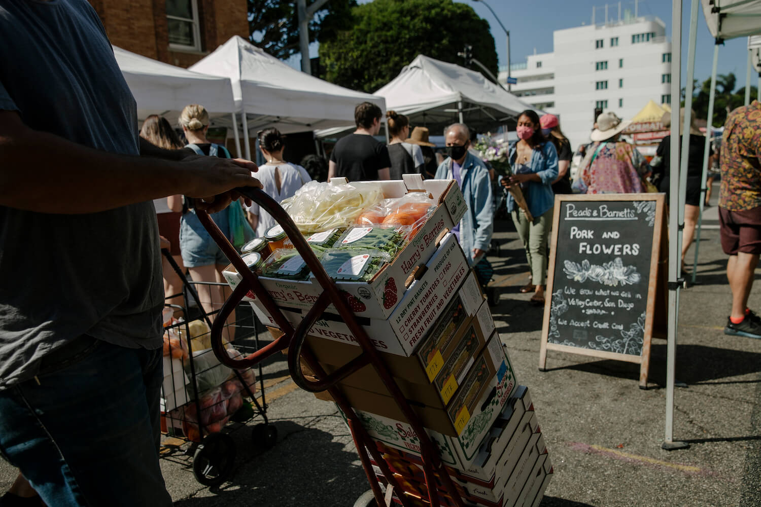 Focused shot on stacked boxes of produce with farm stands and shoppers in the distance at the Santa Monica Farmers Market. August 2021