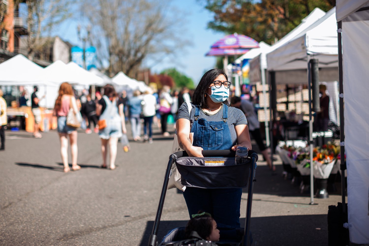 A pregnant mother pushing her little girl who is 1.5 years in a wagon at the local farmers market in Vancouver Washington. She is wearing a face mask while in public outdoors in the covid-19 pandemic.