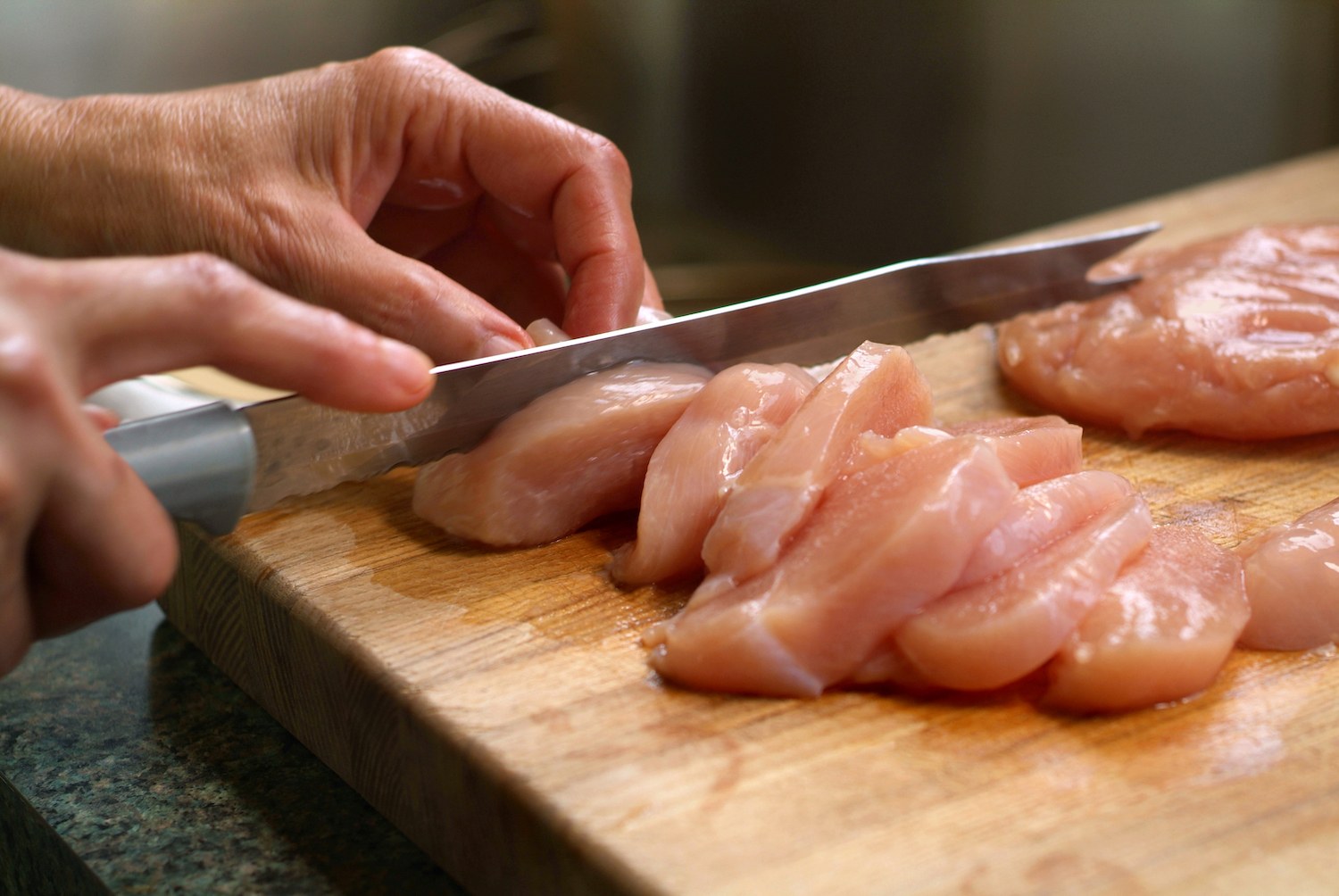 Horizontal image of raw chicken breasts being sliced on a wooden cutting board in a kitchen setting. There is motion blur in the hands and the knife to create a feeling of action, while focus is on the chicken. The photograph was taken with shallow depth of field.