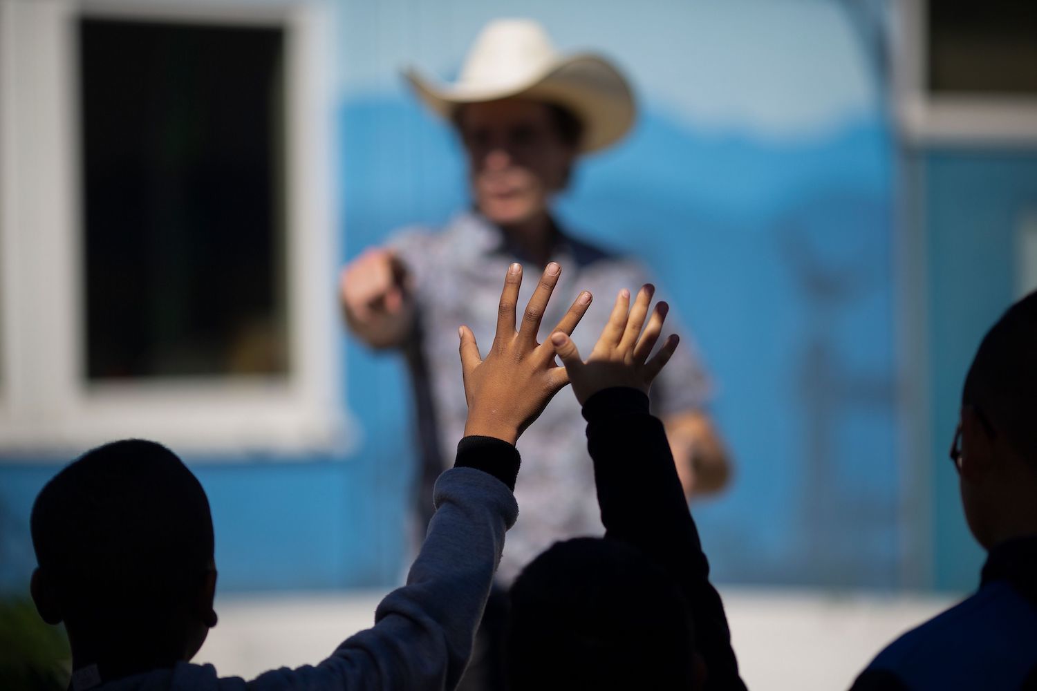 Kimbal Musk blurred in the distance points towards students of color at Eucalyptus Elementary School raising their hands in the foreground. March 13, 2019.