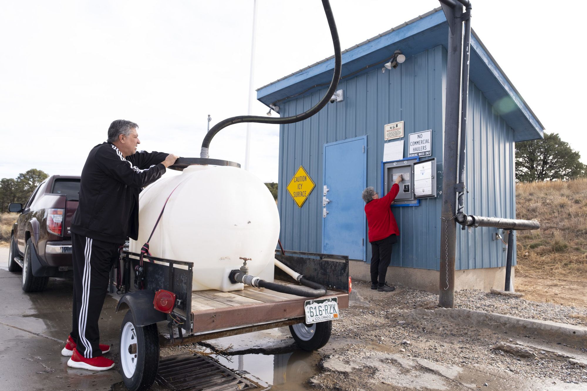 Man in black track suit holds black hose pumping water in white container with little blue shed in background December 2021.
