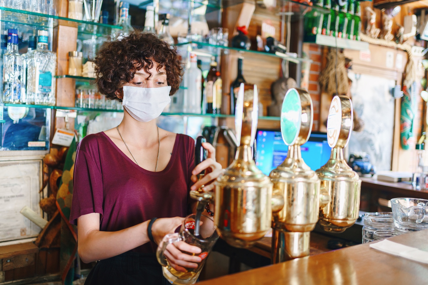 Curly haired masked bartender fills beer. January 2022