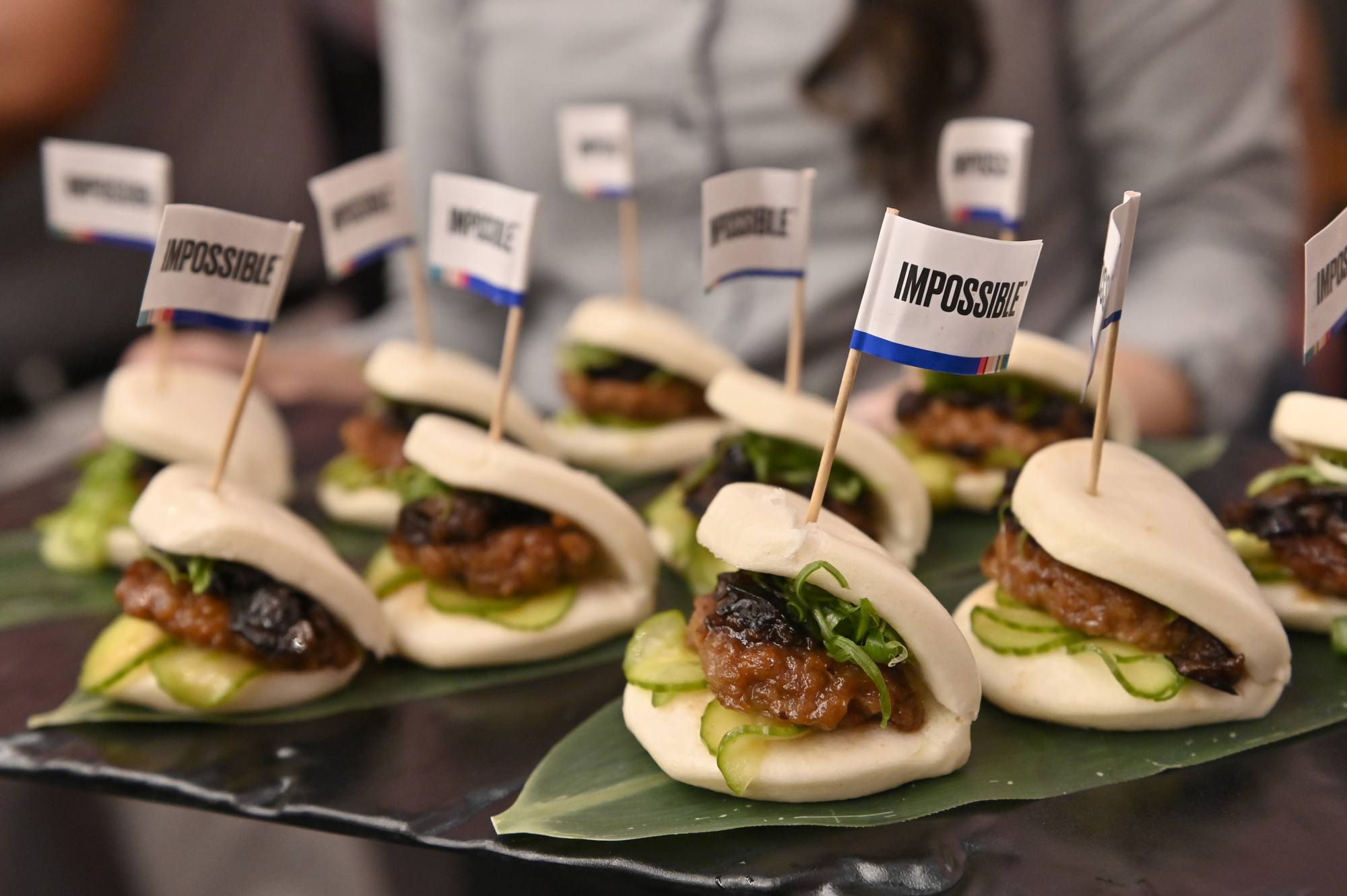 A tray of impossible burgers with pickles, buns, and white impossible flags in them with person in background February 2022.