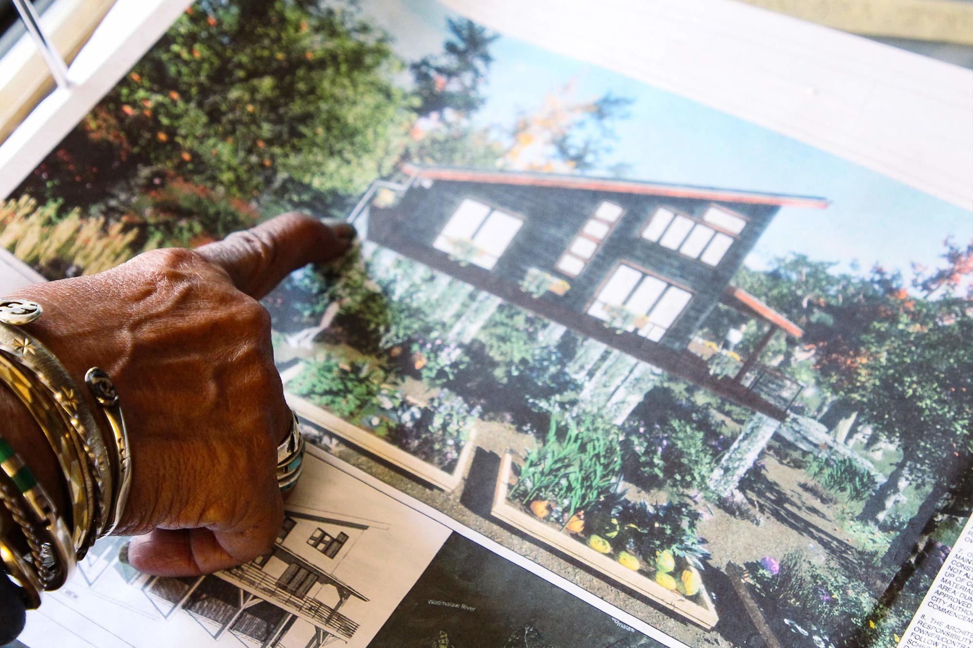 The hand of Arianne King Comer points at a photo of a brown house and green garden with more photos below March 2022.