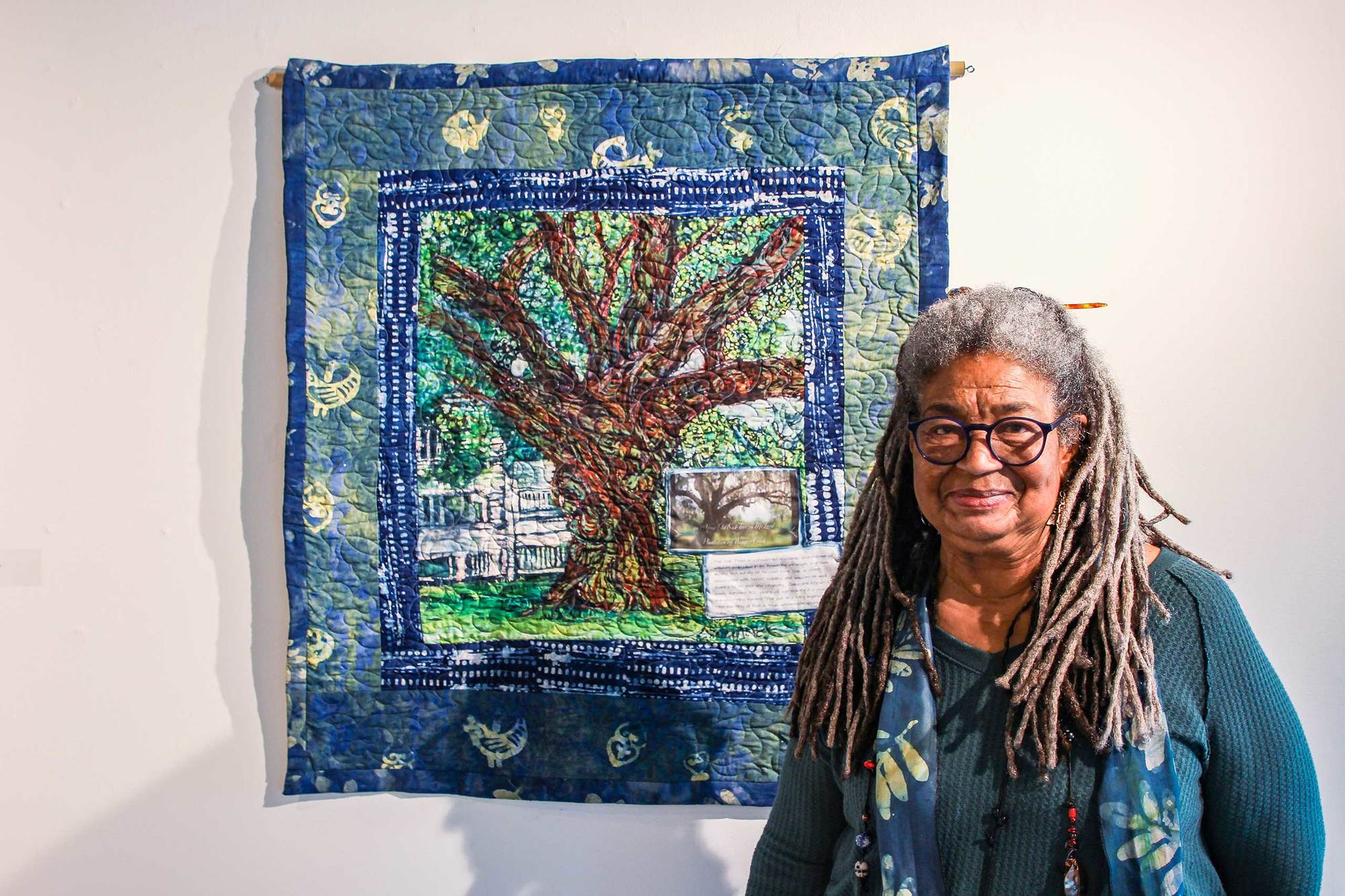 Arianne King Comer smiles at the camera with quilt artwork of tree and blue border with shadow March 2022.