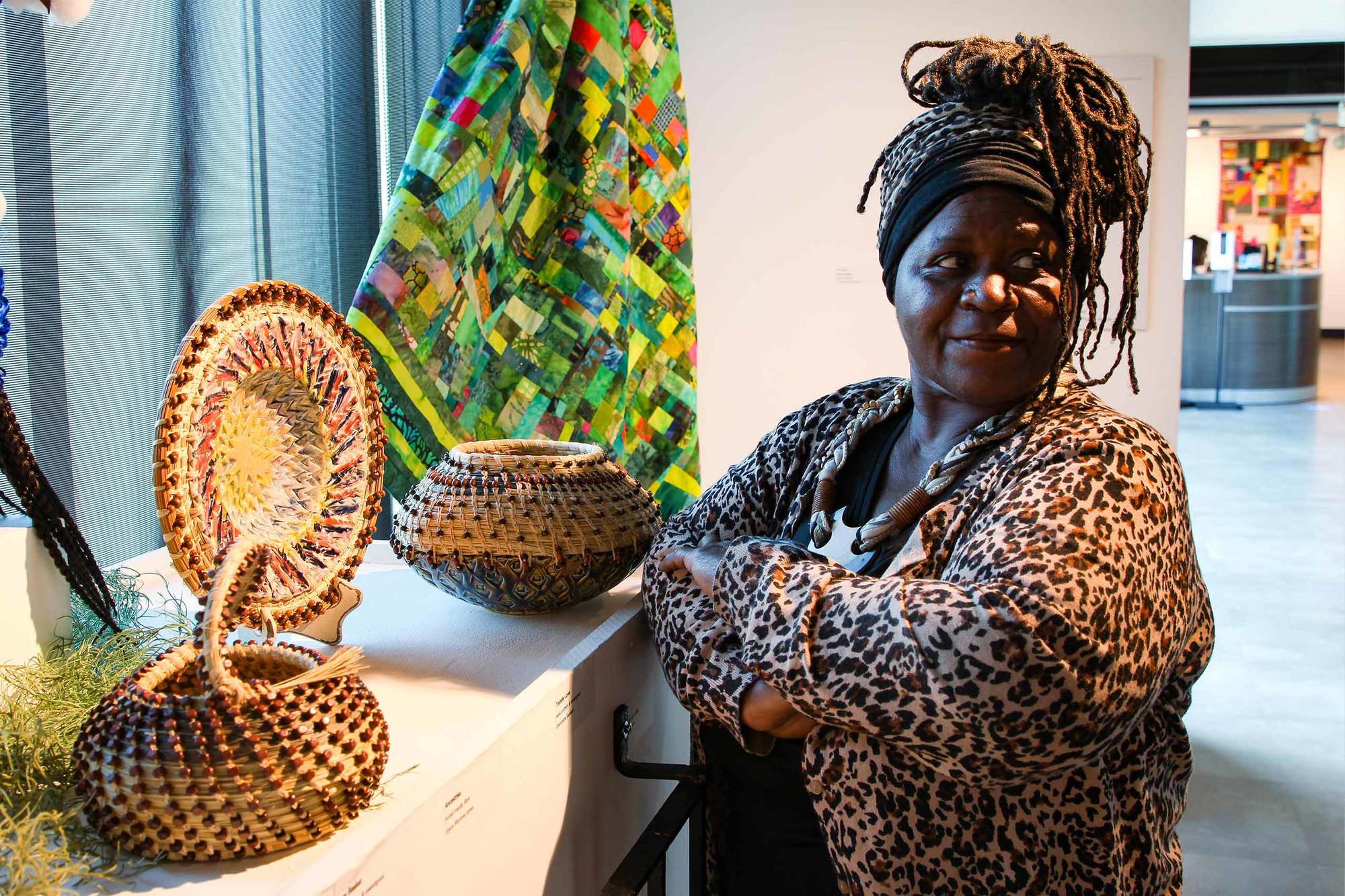 Georgette Sanders crosses her arms and looks away from bowls and art on display with quilt in the background March 2022.