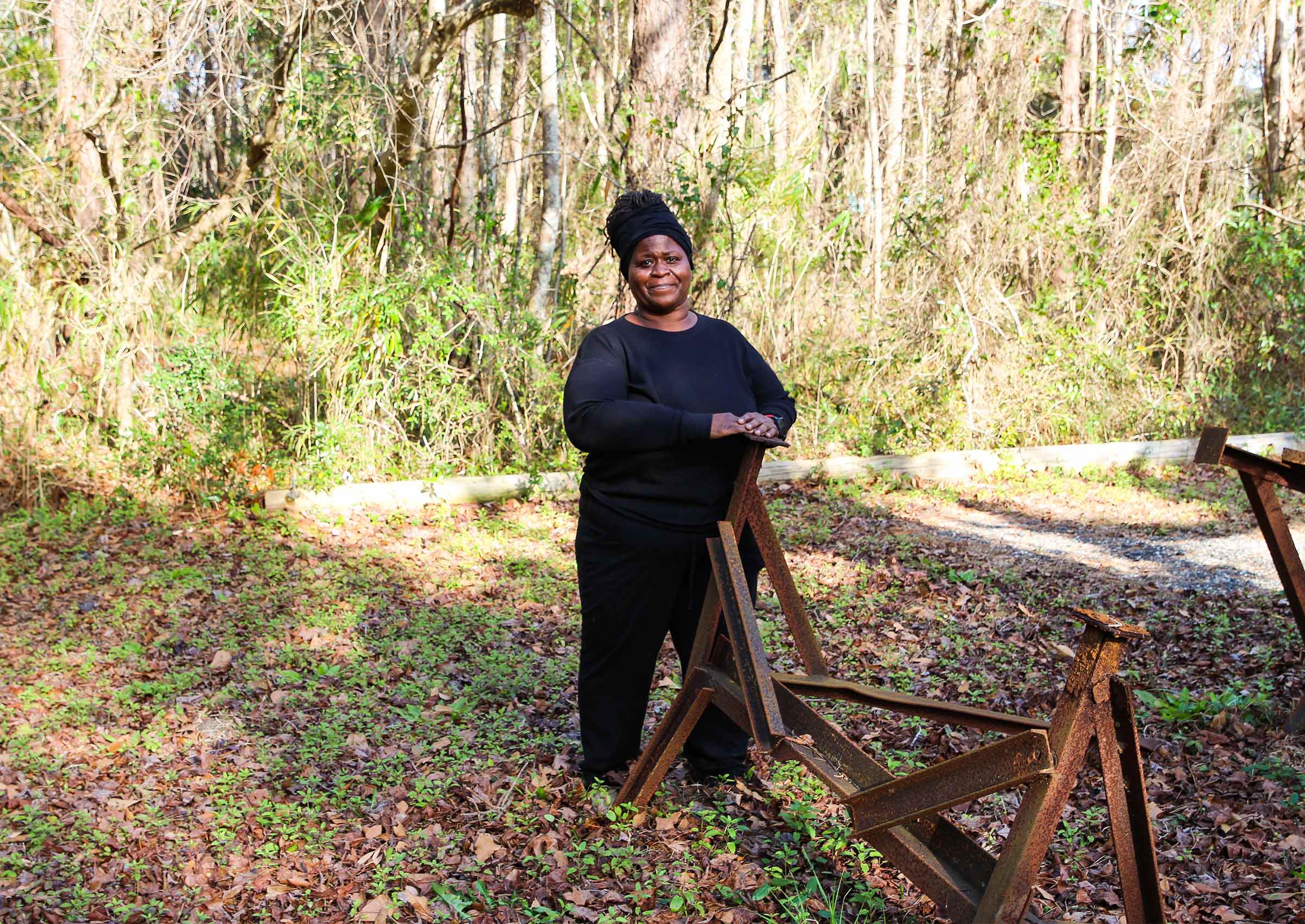 Georgette Sanders poses wearing all black clothes for a camera with her hands placed on a piece of metal and mixed foliage in the background March 2022.