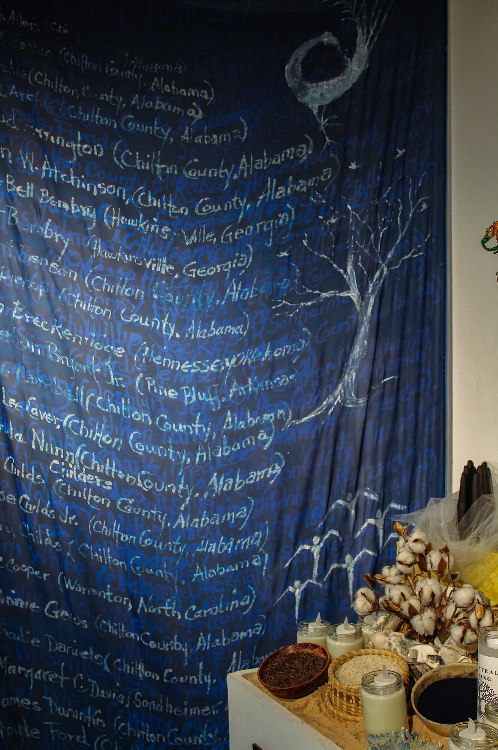 A blue curtain hangs from a wall displaying names along with counties and state names and illustrations of a tree and people March 2022.