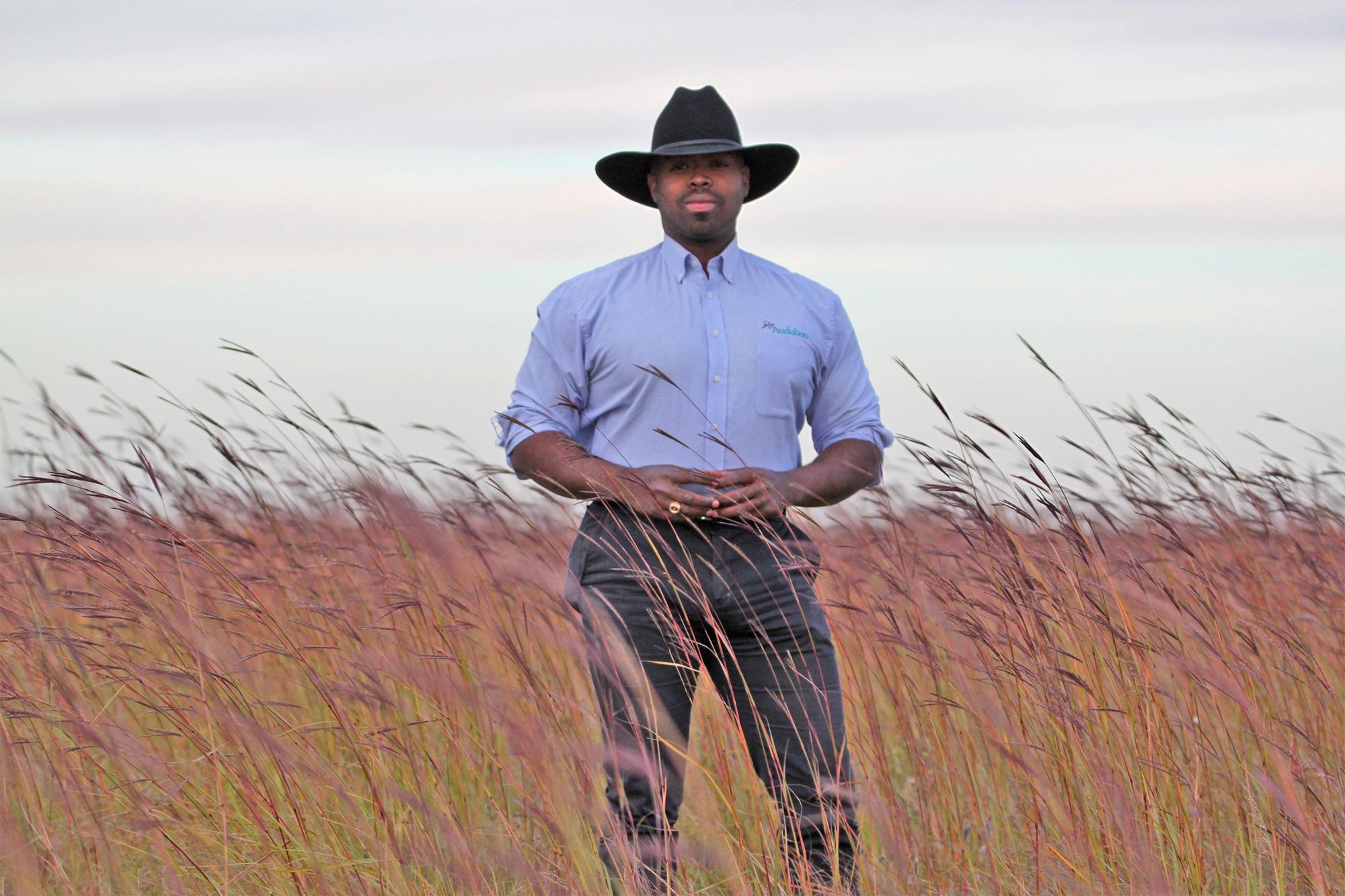 Marshall Johnson stands and looks at camera in field while wearing blue shirt and black cowboy hat April 2022.